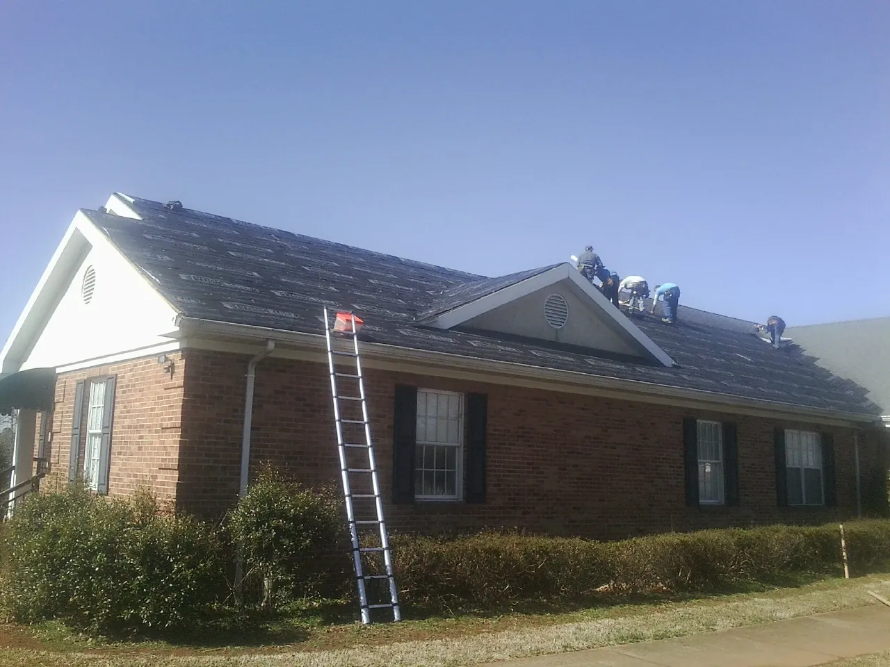 Crew tearing shingles off a brick ranch roof with a ladder set — Emergency Roof Repair work by our crew in Henrietta